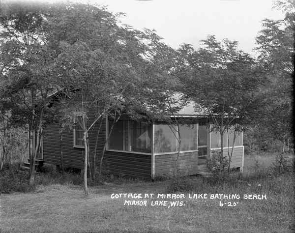 Three-quarter view of a small cottage at Mirror Lake Bathing Beach, with a screened-in porch and small trees in the yard.