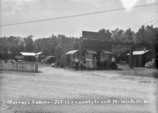 View across road towards a group of people standing in front of the main office and general store at Murray's Cabins. On the right is a wooden archway with an American flag that reads: "Main Entrance, Cabins, Trailers." There are signs for Rothe's Ice Cream and Braumeister Beer near the store.