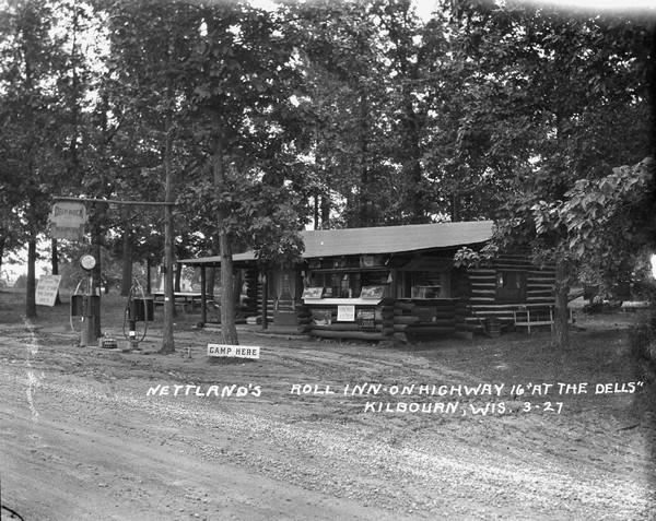 View across road towards the main office at Nettland's Roll Inn. The building is of chink log construction with the round logs overlapping at the ends. Candy, soda and ice cream are for sale at an open counter in front. Two gas pumps stand at the side of the road among trees.
