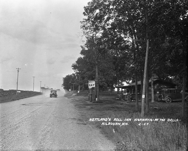 View down road with the Nettland's Roll Inn on the right. An automobile is coming down the dusty road towards the inn, which has an "EAT" sign handing over the gas pumps near the side of the road.