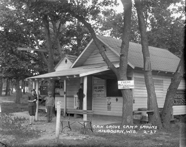 Four young people standing in front of the main office and general store at the Oak Grove Campground. A sign on a tree reads: "Fresh milk, cream, eggs and butter."