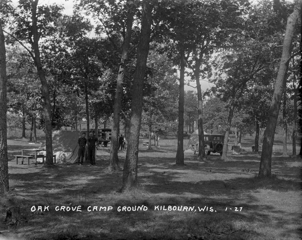 View of the camp sites under the trees at Oak Grove Campground. Two men stand near a tent on the left.