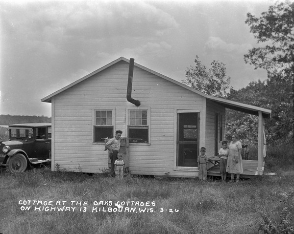 Family of five standing in front of a small guest cottage with porch. There is an automobile parked on the left.