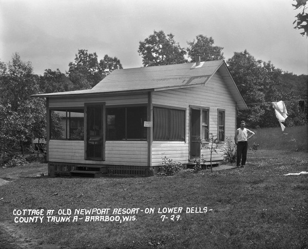 Exterior view of a small guest cottage with screened porch. A sign on the side of the cottage reads: "Minnows for Sale," and a man is standing under the laundry line on the right.