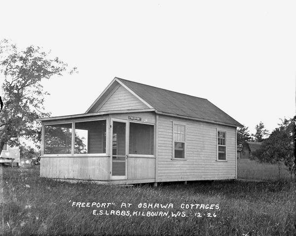 The "Freeport" cottage at Oshawa Cottages. There is an entrance on the side of the screened-in porch. In the background on the left is another building, and what appears to be a parked automobile.
