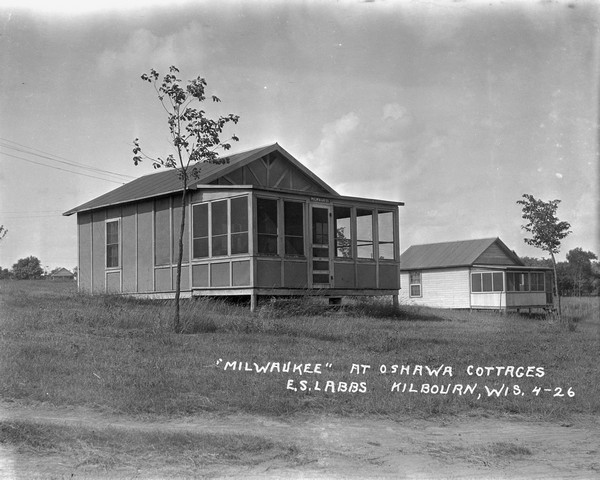 Exterior view of the "Milwaukee" guest cottage at Oshawa Cottages. There is a screened-in porch with steps to the door. Young trees are planted in the yard, and another cottage is in the background on the right.