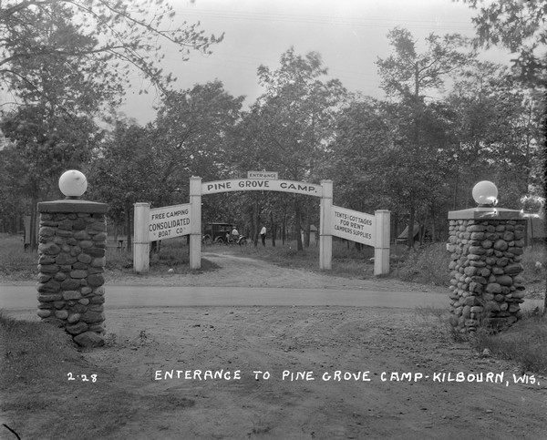 View from driveway with stone columns, over road towards the entrance gate to the Pine Grove Camp. People are standing near an automobile, tents and an outhouse in the background under the trees.