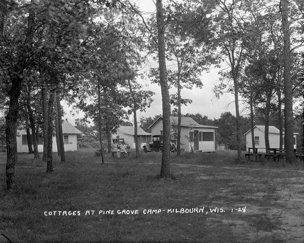 View of a group of guest cottages at Pine Grove Camp. Automobiles are parked by two of the cottages. A woman and a girl are sitting on a park bench under the trees.