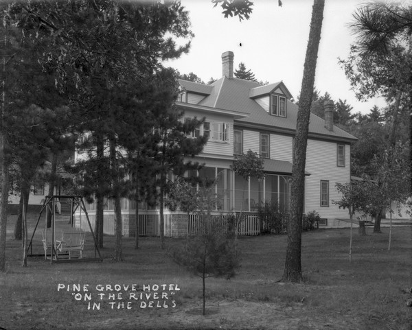 View across lawn of the Pine Grove Hotel, a two to three-story building with a large, wrap-around porch. There are pine trees and a lawn swing in the yard. Another building is in the background on the left, and a garage or other outbuilding is in the background on the right.