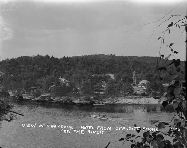 Elevated view of the Pine Grove Hotel and Resort from the opposite shore of the Wisconsin River. An excursion boat is passing by. Rock formations are along the shoreline.