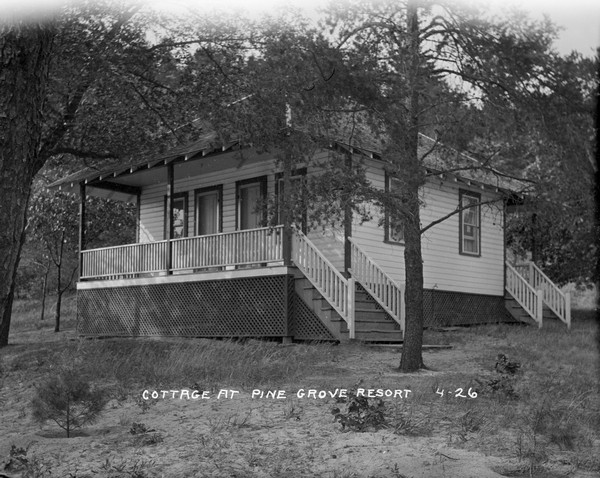 View of an elevated guest cottage on the slope of a hill with two entrances, one at the front with steps leading up to a porch, and one at the back, at Pine Grove Resort. The yard is sandy and has patchy grass.