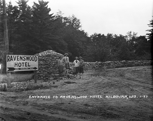View of the entrance drive of the Ravenswood Hotel. A group of four women are standing next to the stone wall and sign at the driveway off the road. All the women wear bloomer pants and patterned socks, and two of the women are wearing middy blouses.