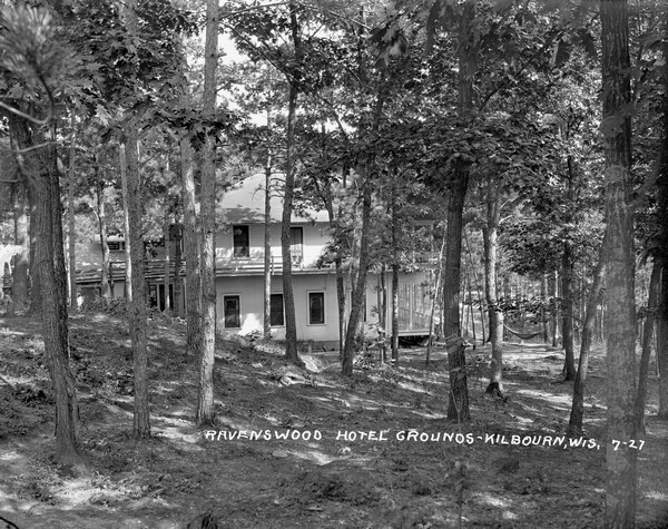 View down slope of hill towards the side of the Ravenswood Hotel among the pines. There is a porch and balcony along the side and front of the hotel, and hammocks are set up among trees on the right.