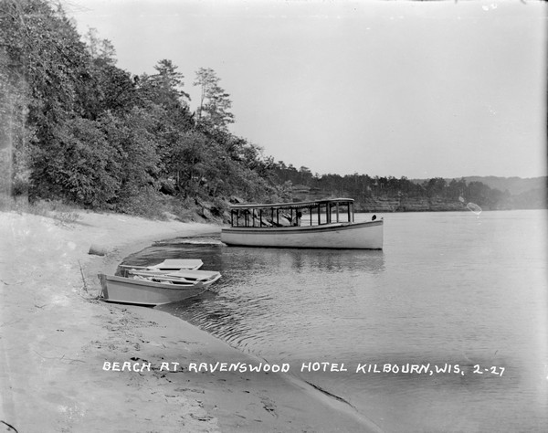 The Ravenswood beach on the Wisconsin River. A man is piloting an excursion boat as it is pulling away from the shoreline. Two rowboats are pulled up on the shore. Rock formations and cliffs are in the background.