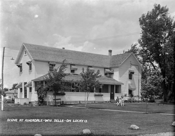 The main building at Riverdale Resort. Three children are sitting on a bench on the lawn. Awnings are on all the windows, and there is a screened-in porch.