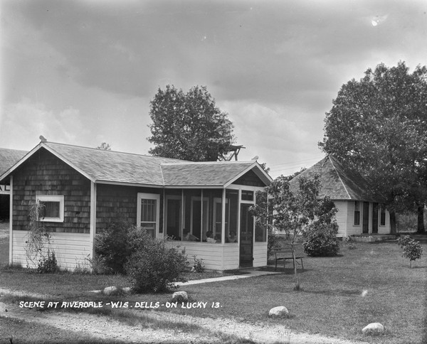 Two cottages at Riverdale Resort. There is a group of guests in the screened-in porch in the cottage on the left. Behind the tree in the center is a water tower.