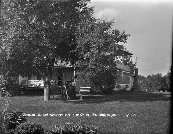 Three-quarter view of Glen Rood's Resort. A  man, woman and child are near the lawn swing in the yard. Benches are on the open porch and in the yard. There is a balcony above the porch.