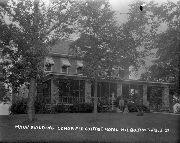 The main building of Schofield Cottage Hotel with a large porch and stone columns. On the lawn in front posing is a man standing, and two children sitting, on the lawn. A woman is standing near a column on the right under the Schofield Hotel sign, and on the lawn behind her is a lawn swing.