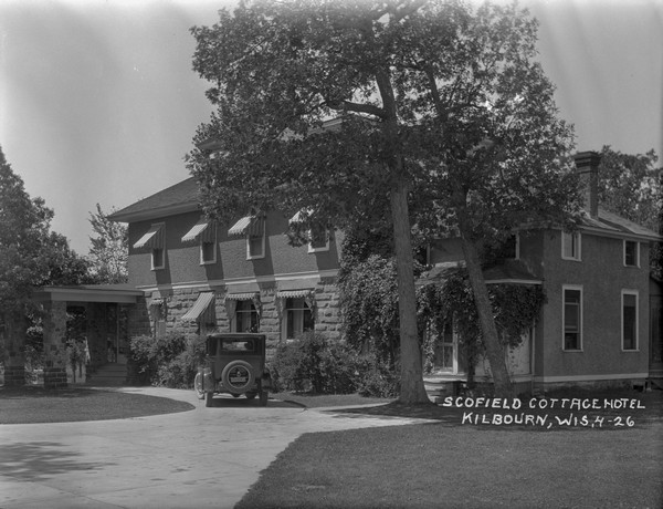 Schofield Cottage Hotel and driveway with an automobile parked near the porte-cochere. There are striped awnings over the windows on both the first and second floors. Heavy vines cover the screened-porch entrance on the right side of the hotel.