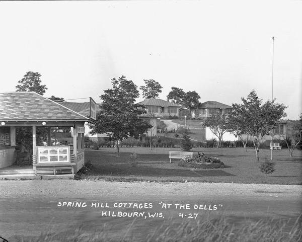 The grounds at Springhill Cottages, with four cottages on the hill in the background. There is a person sitting on a chair inside an open-sided building in the foreground on the left, which is selling postcards and tickets to a tour boat line. More people sit on benches on the lawn in the background.