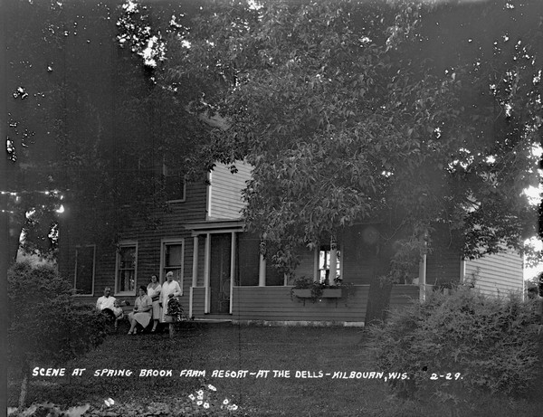 A group of people are gathered on and around a bench on the lawn in front of the porch.