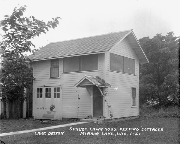 Two-story cottage with a built-in garage. There is a young vine over the roofed entrance, and a bench and potted plant near the garage door.