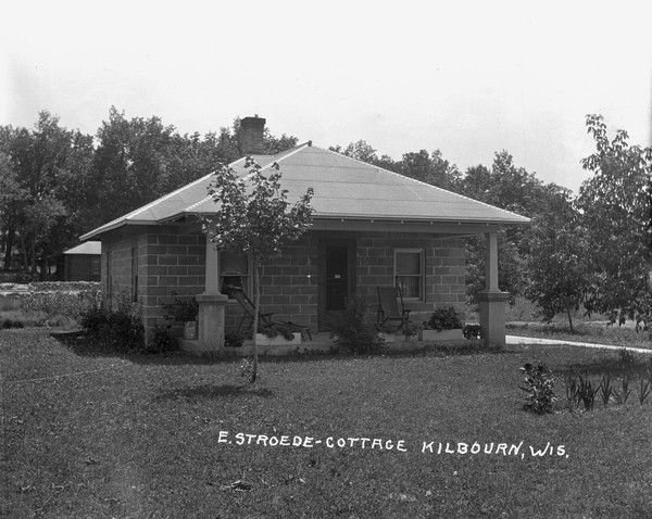 Exterior view of small cottage made of cement blocks. There are convertible lounge chairs and flower boxes on the concrete porch. There is another building in the background near trees.