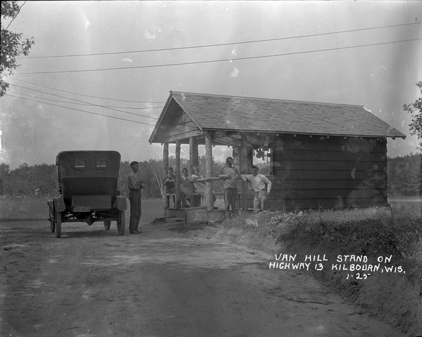 Vending stand on Highway 13 near Van Hill Cabins. A man stands near an automobile parked in the road in front, and three girls are gathered on benches under the porch. Two younger men stand along the railing. A small sandwich board near the road reads: "Tri-State Ice Cream."