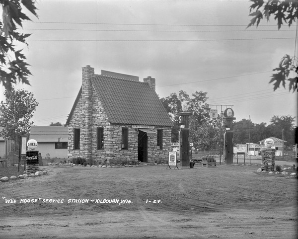 Exterior view of a stone building which has a steep, tiled roof, and an awning over the open doorway under the "Wee Hoose" sign. In front are pumps for Shell gasoline, and behind on the left are cottages for rent. The White House Hotel is across the road on the right.