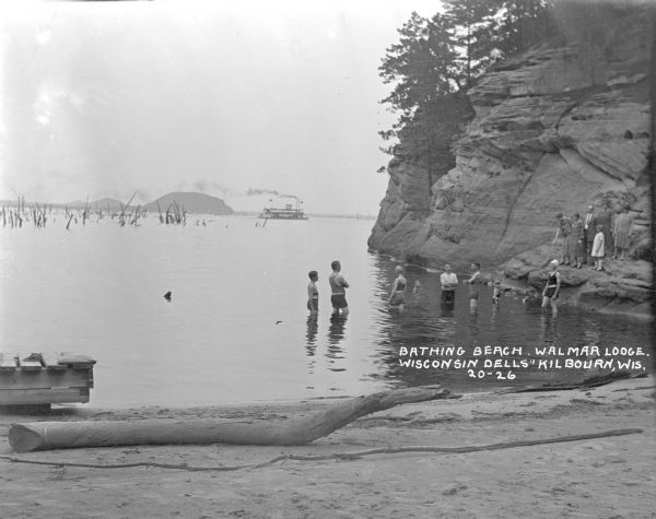 Beach on the Wisconsin River. There is a group of bathers in the water, and a group of tourists on a nearby rock. A steamboat is passing by in the distance.