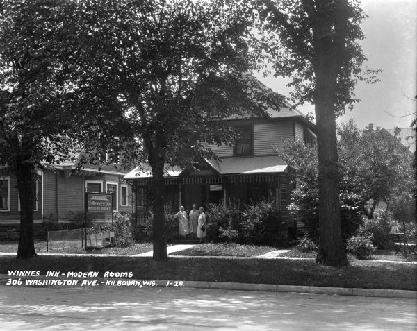 View across street towards three women standing on the front steps of Winnes Inn, a converted dwelling in a residential neighborhood.