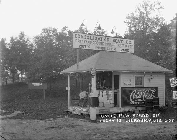 Roadside kiosk that provides information and sells candy, soda, gasoline and tickets for Dells attractions. A man is sitting on the porch, and pennants hang from the counter.