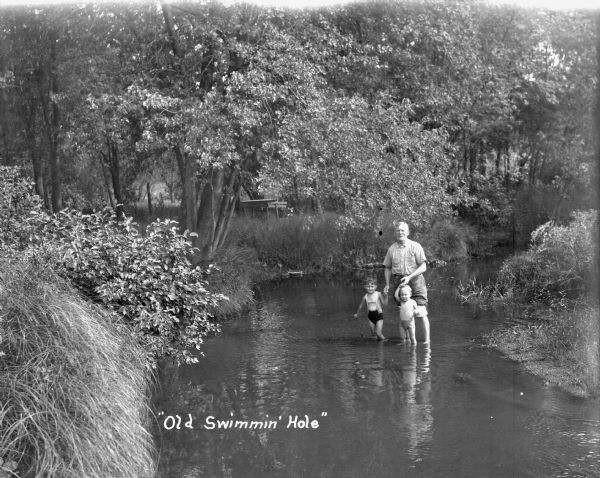 A man holds the hands of two children, all posing standing in the middle of a stream. The two children wear swimming suits, and the man has his pants rolled up. Trees and bushes are along both sides of the stream bank.