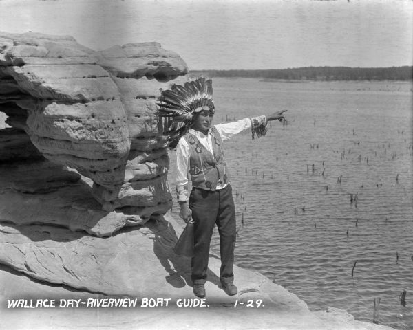 Native American tour guide standing on a rock formation. He is wearing a headdress and decorated vest and white shirt, and is holding a megaphone and pointing towards the river.
