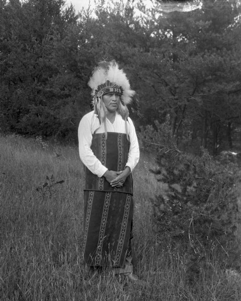 Full-length portrait of a Native American man standing in tall grass near trees. He is wearing headdress and an Indian blanket.