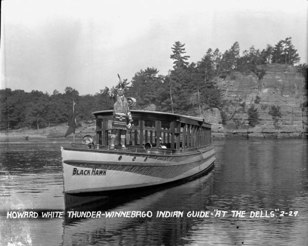 Native American guide in native dress standing on the bow of a tour boat floating in the river. A pilot wearing a hat is sitting behind him. On the front of the tour boat is a painted sign reading: "Black Hawk." Rock formations are on the far side of the river.