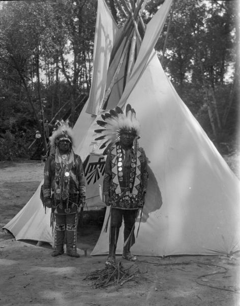 Portrait of two Native American men wearing feathers and beadwork who are standing in front of a teepee, which has a thunderbird symbol inside, seen through the open front doorway.