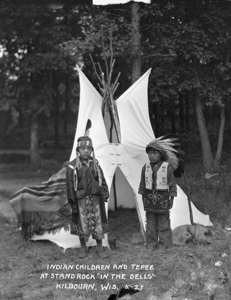 Two Native American children, a girl and a boy, standing in front of a small teepee with a blanket draped over part of it.