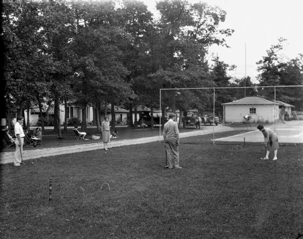 People playing croquet on a lawn next to tennis courts. In the background on the left, people are relaxing under trees across the road. Cottages are in the background.
