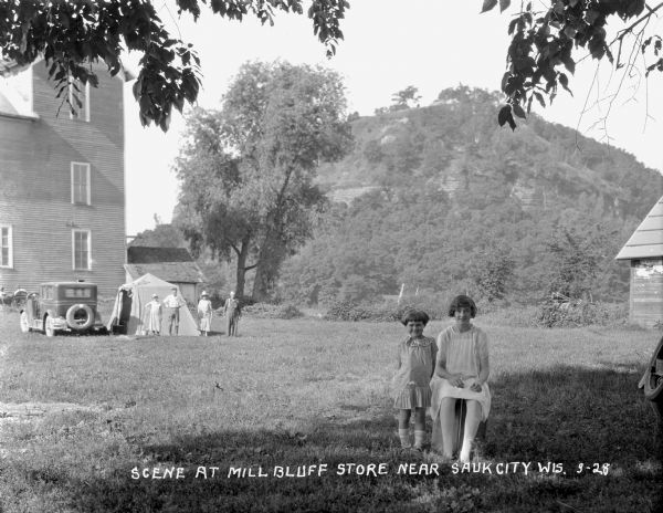 Two girls standing and sitting in the foreground on a lawn. In the background on the left a group of people stand near a tent and a parked automobile. Behind them is a three-story wood frame building. In the background is a tree-covered bluff.