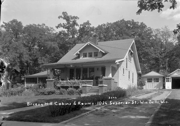 Three-quarter view of front of house with two garages in the back. There are bushes along the driveway and flowers in the box along the front of the porch.