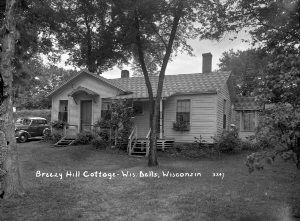 Duplex cottage with automobiles parked in the back and on the left. Flowers are in window boxes.