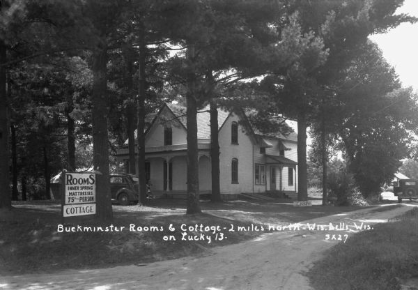 View down driveway towards main house of Buckminster Rooms. A sign in front advertises rooms with inner spring mattresses for 75 cents per person. An automobile is parked in the drive and near the front porch.