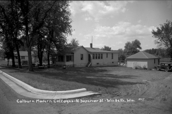 Main house for Colburn's Modern Cottages on the Wisconsin River. A curb is in the foreground, and a dirt road leads to a garage and a cottage. An automobile is parked next to the garage. Patio furniture is on the lawn.