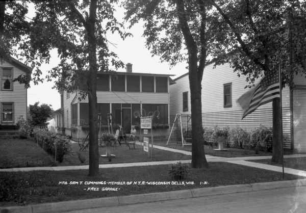 View from street of a couple is sitting on the front stoop of a house which has rooms for tourists. A lawn swing and planters are on the lawn. There is a large flag on a flagpole flying at the curb.