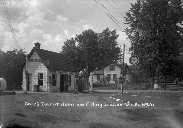 The filling station and main office at Drew's Tourist Home. There are flags on the gas pumps. A man and his dog are standing next to the building.