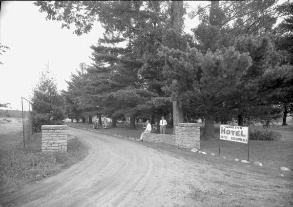 View of the entrance drive to the Grand View Hotel with stone columns and stones framing the drive. Two men are posed near the right columns, and a three people pose further down the drive near chairs and a lawn swing on the lawn. Large pine trees obscure the hotel.