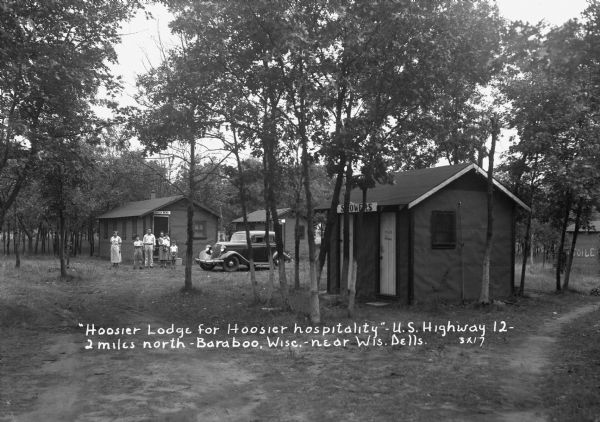 View down dirt road of the tourist camp with cottages. There is a building for men's and women's showers in the foreground, and a building marked "Toilet" is in the background on the right. On the left is a group of people standing near a cottage called "South Bend." An automobile is parked in front, and another cottage is next door.