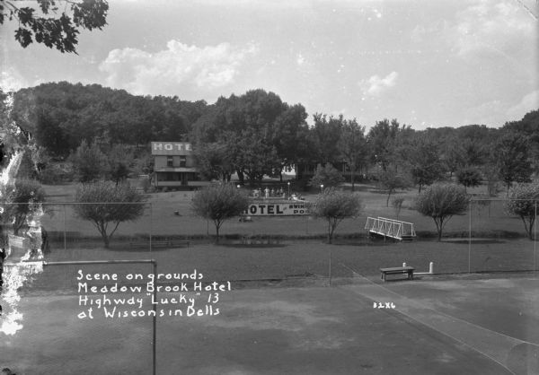 Elevated view over the volleyball court of the grounds at the Meadow Brook Hotel. A footbridge is across a stream on the right. A group of people are standing by the above ground swimming pool on the other side of the stream, and the hotel's main building and annex is behind them in the background. A wooded hill is behind the hotel.