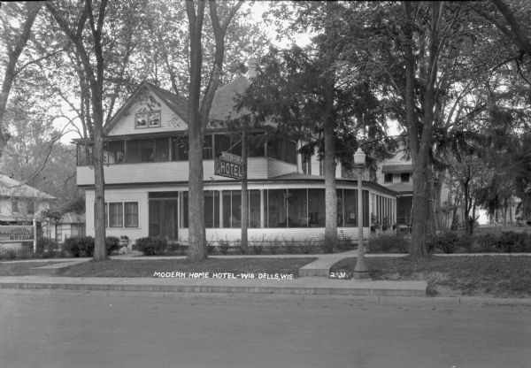 Exterior view across street of the Modern Home Hotel in a residential neighborhood. There are large, enclosed porches on the first and second stories. A lawn swing is in the yard on the right.